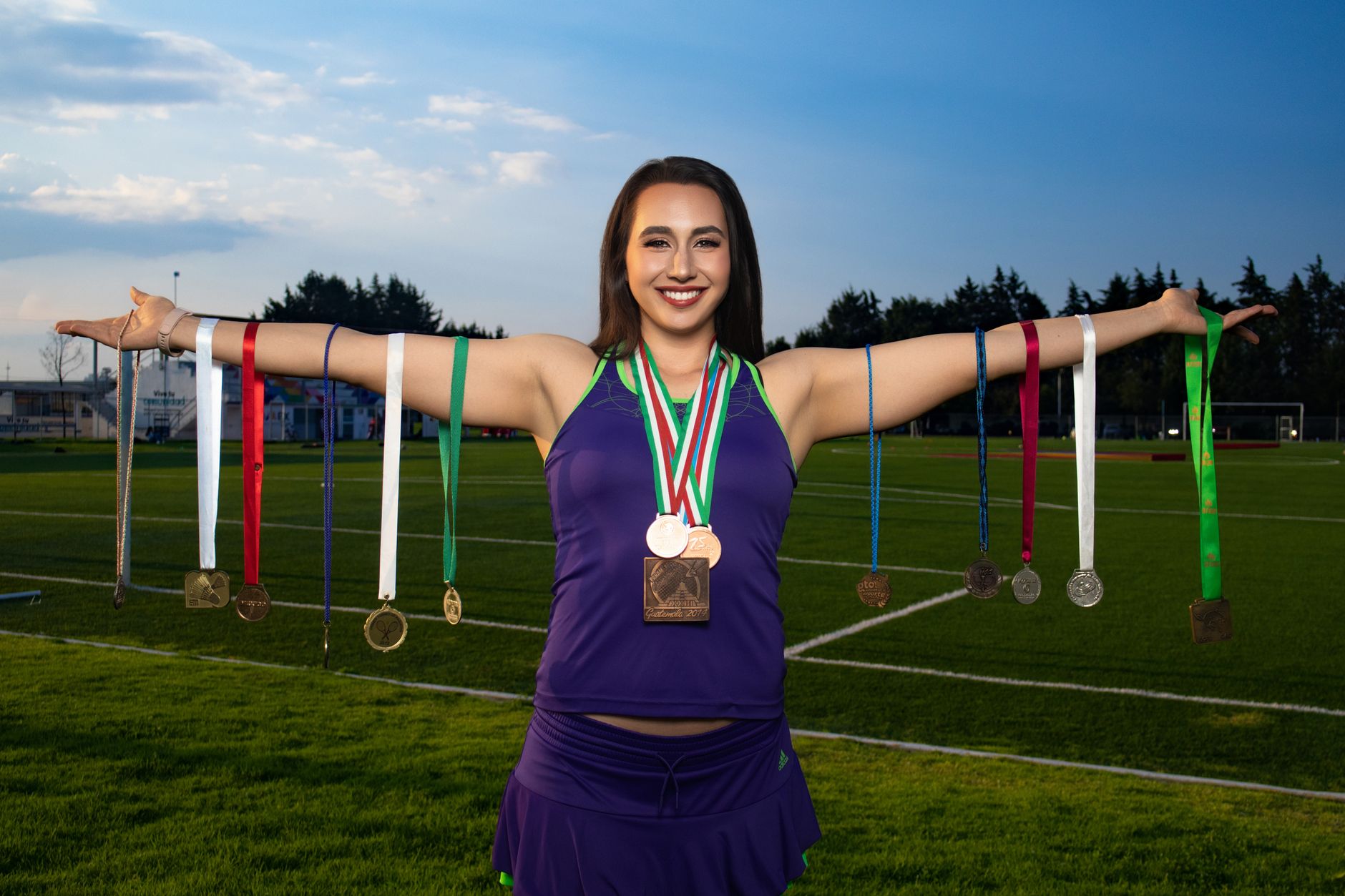 Smiling female athlete holding numerous medals on an outdoor sports field.