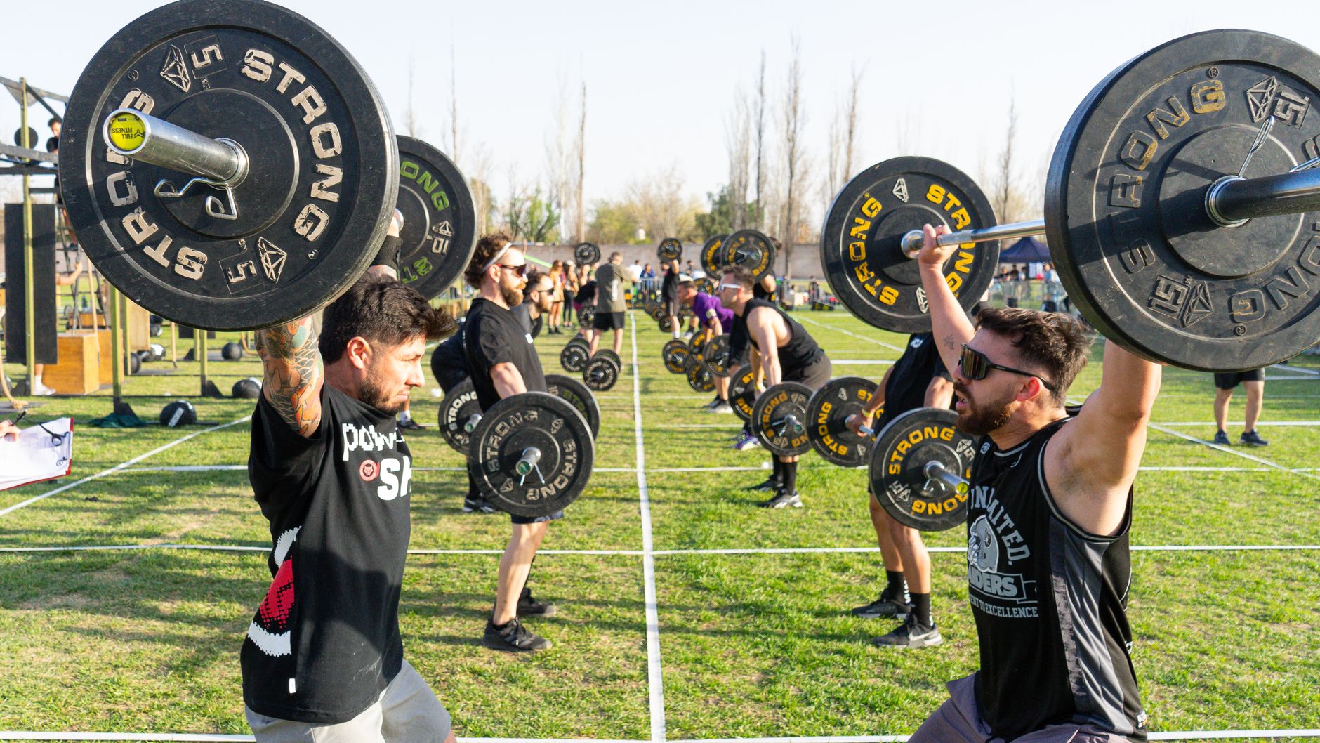 Strong athletes competing in an outdoor weightlifting event, lifting barbells in daylight.