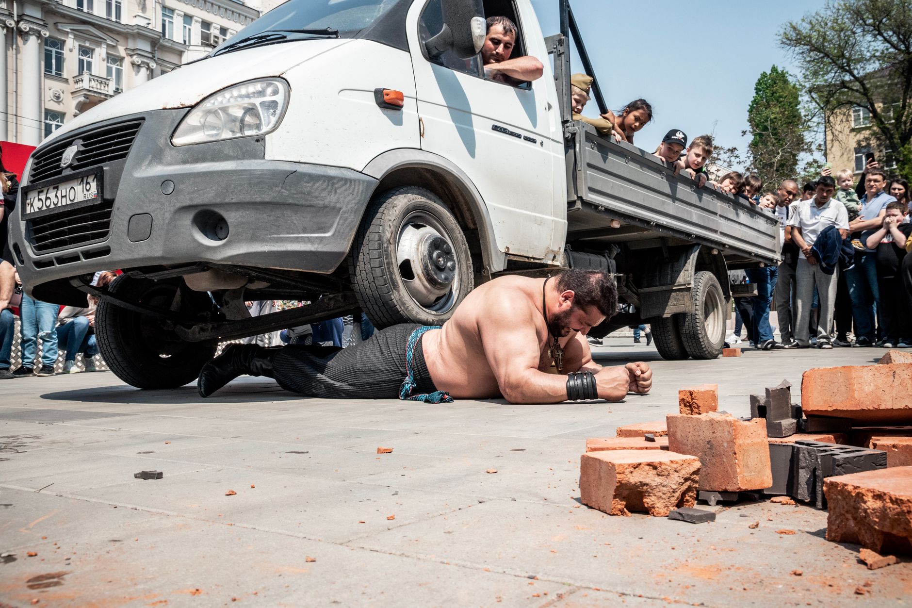 A strongman performs a stunning feat by lifting a truck while a crowd watches outdoors.
