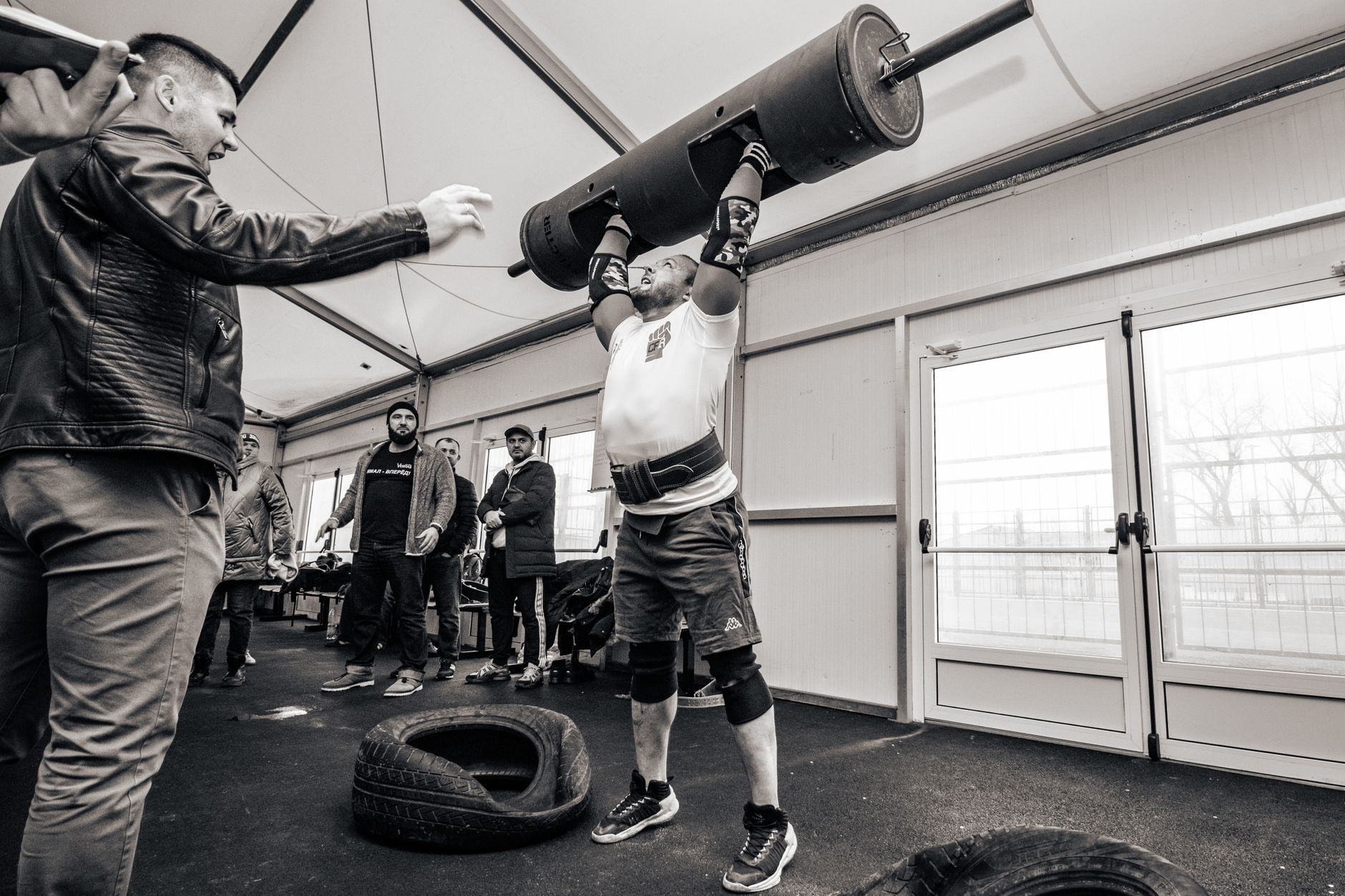 A strongman lifts a barbell overhead during an indoor gym event, showcasing strength and determination.