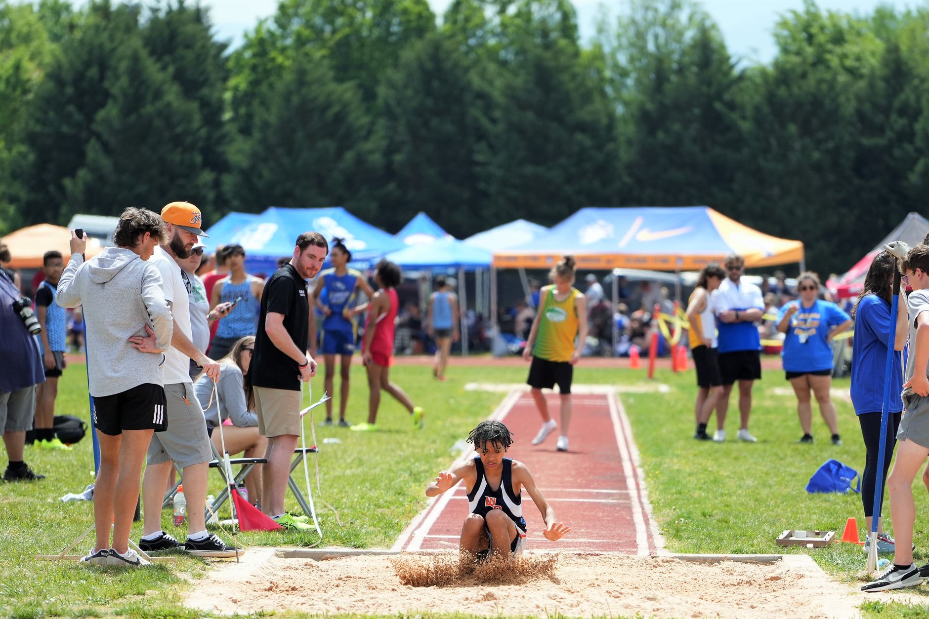A teenage athlete performs a long jump during a track and field competition on a sunny day.